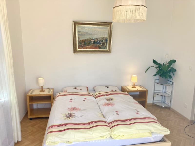 The image shows a cozy bedroom with a double bed, two nightstands, a framed painting on the wall, and a potted plant. The bed is made with a floral patterned comforter and the room has a warm, inviting atmosphere.