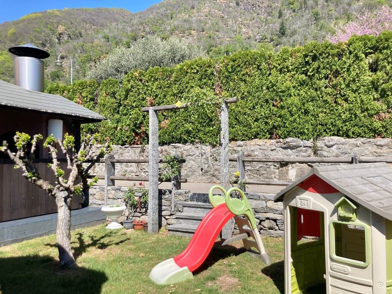 Outdoor area with a stone wall, a slide for children, a small playhouse, and a chimney. The area is surrounded by lush greenery and mountains in the background.