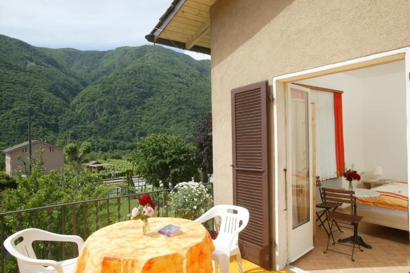Apartment with a balcony overlooking a lush, green mountainous landscape. The balcony has a small table and chairs, and the interior of the apartment is visible through the open glass doors.