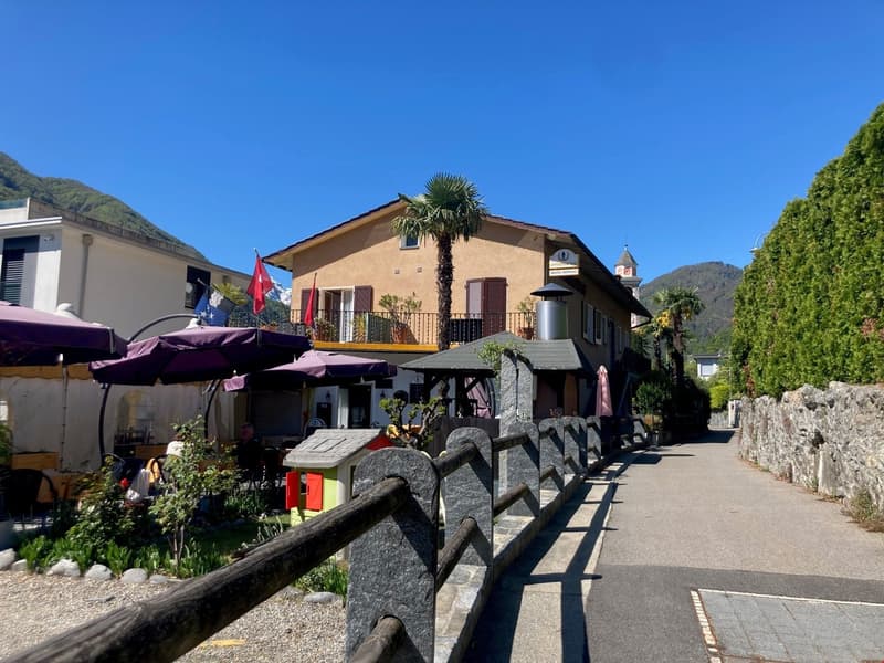 Two-story building with a Mediterranean-style architecture, featuring a balcony and a palm tree on the roof. The building has a restaurant or cafe on the ground floor, with outdoor seating and umbrellas. The building is surrounded by greenery and mountain