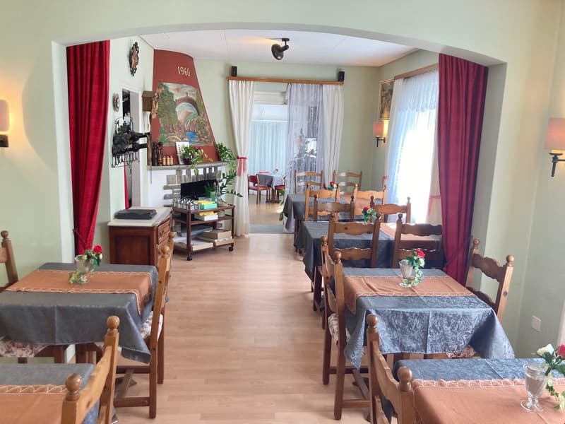 The image shows a cozy dining room with wooden furniture, including tables and chairs. The room has a fireplace and decorative elements like wall art and potted plants. The space appears to have a warm and inviting atmosphere.