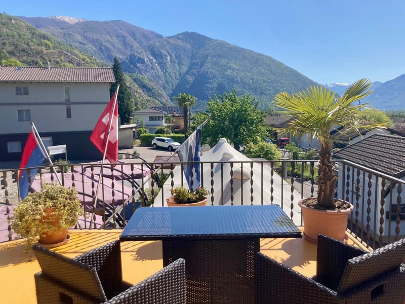 The image shows an outdoor patio or balcony area with a table and chairs, surrounded by lush greenery and mountains in the background. There is a Swiss flag visible, indicating this is likely a location in Switzerland. The patio has a railing and appears