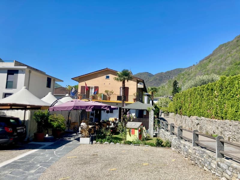 A two-story building with a terrace, surrounded by lush greenery and mountains in the background. The building has a Mediterranean-style architecture with a tiled roof and a palm tree on the balcony. There is a seating area with umbrellas in the front of