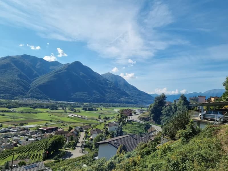 The image shows a scenic mountain landscape with a village nestled in the valley below. The mountains in the background are tall and rugged, with a blue sky dotted with clouds overhead. The village consists of a cluster of buildings with red-tiled roofs,