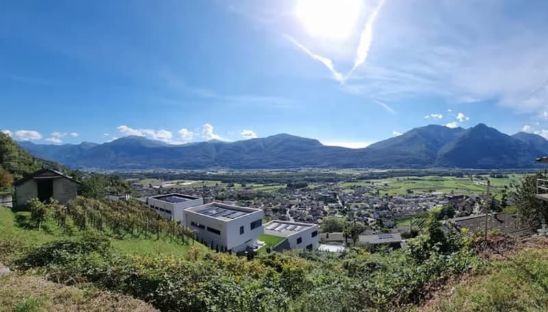 This image shows a panoramic view of a town or city nestled in a valley surrounded by mountains. The landscape features rolling hills, green vegetation, and a clear blue sky with some clouds. There are several modern-looking buildings visible in the foreg