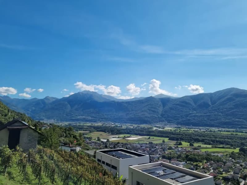 The image shows a panoramic view of a mountainous landscape. In the foreground, there are some buildings and a solar panel installation. The background is dominated by a range of rugged, snow-capped mountains under a blue sky with scattered clouds. The sc