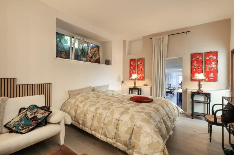 White walls, wooden floor, queen size bed, decorative pillows, two lamps, side tables, shelves, open window, red wall paintings, draped window, oriental decorations