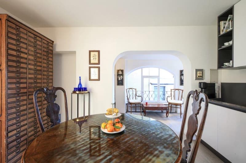 Dining area with wooden table and chairs, large arch window, wooden filing cabinet, kitchen with coffee machine and modern cabinets, small round table with chairs, picture frames on wall