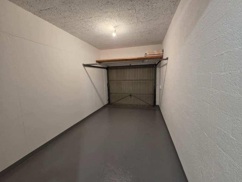 Empty garage with white walls, gray floor, and a wooden shelf above the garage door