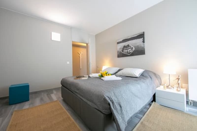 large bed, gray sheets, beige carpets, bedside table with lamp, blue stool, decorative elements