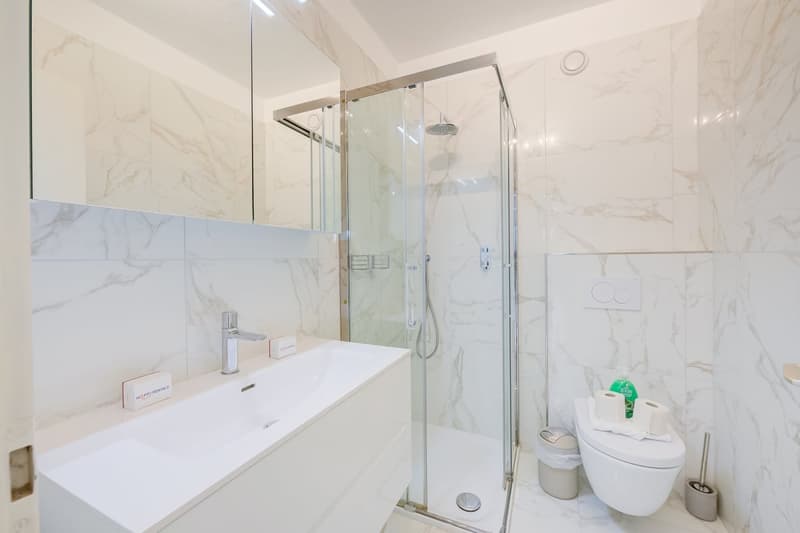 marble walls, white countertop, shower with glass door, sink, faucet, toilet, toilet paper, trash can, toilet brush