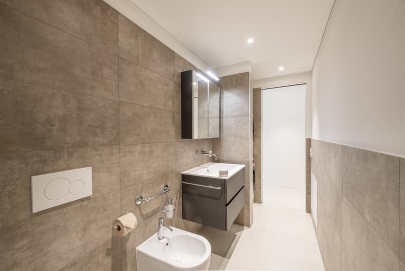 Tiled walls, white floor, toilet, bidet, vanity with sink and storage drawers, mirror, toilet paper holder, and a hand soap dispenser.