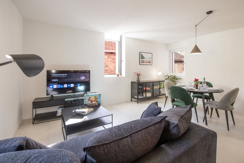 White walls, floor lamp, TV on entertainment center, dining table with chairs, picture frame, coffee table with books, green chairs, ceiling lamps, small window with outdoor view.