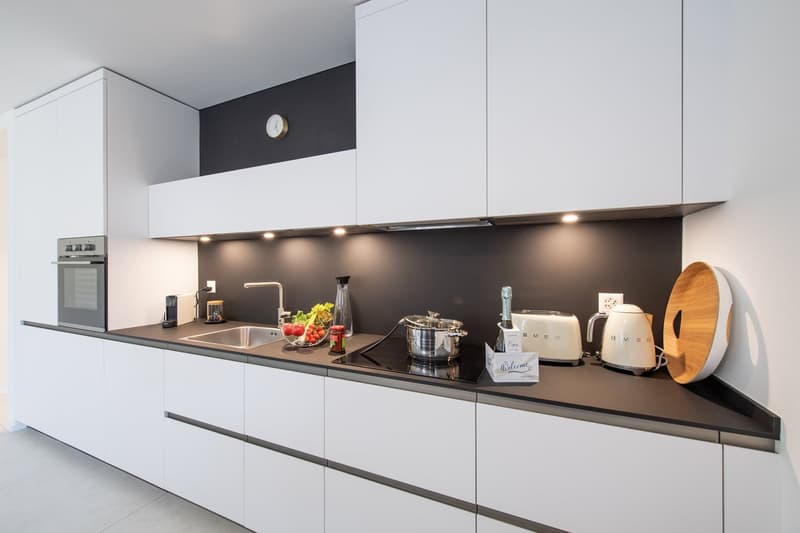 modern white kitchen with black countertops, integrated oven, black induction stove, sink, toaster, kettle, wooden chopping board, under cabinet lighting