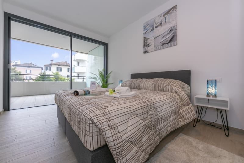 modern bedroom with wooden floor, white walls, gray headboard bed, plaid bedding, nightstand with lamp, sliding glass door leading to balcony, greenery view outside
