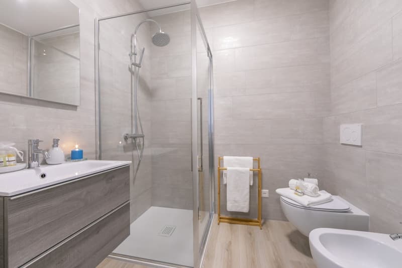 Modern bathroom with glass shower enclosure, white tiles, wooden floor, vanity with sink, toilet, bidet, towel rack, and towels