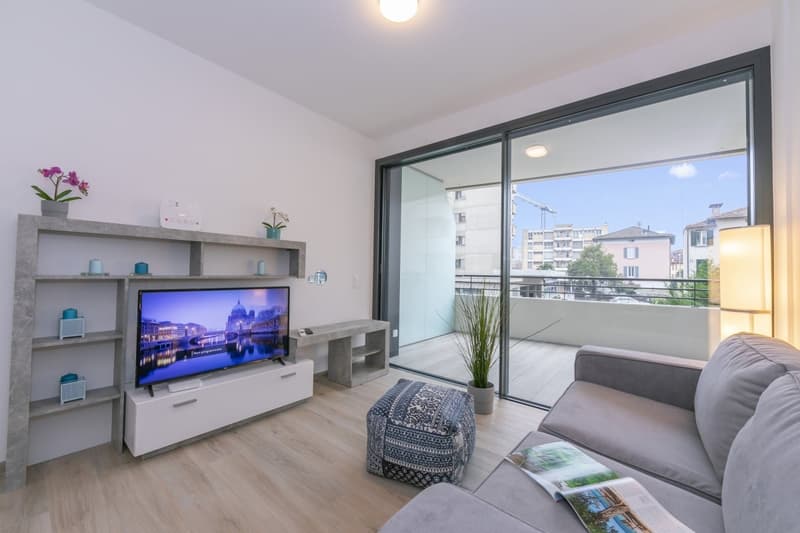 open living room with tv on concrete stand, gray sofa, ottoman, sliding doors to balcony, glass windows, hardwood floor, plants, lamp, cityscape view