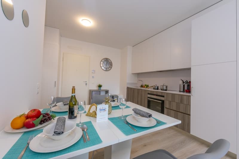 modern white interior, dining table, chairs, wooden floor, white cabinets, kitchen area, fruit plate, wine bottle, plates, glasses, forks, spoons, napkins