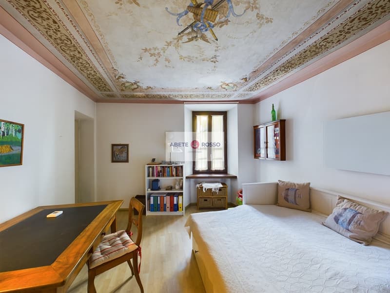 Spacious living room with ornate, painted ceiling, wooden furniture including a desk and shelving unit, and a cozy seating area with a sofa and pillows.