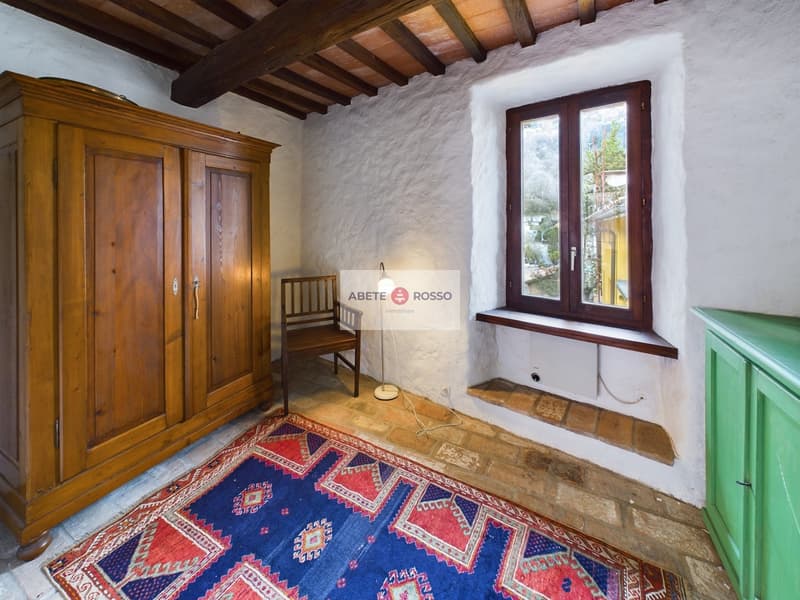 Rustic bedroom with wooden beamed ceiling, large wooden armoire, antique chair, and colorful patterned rug on the floor. The room has a cozy, traditional feel with the exposed stone walls and wooden accents.
