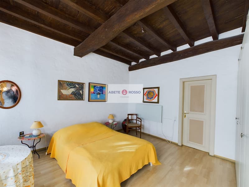 Spacious bedroom with wooden beamed ceiling, white walls, hardwood floor, yellow bedspread, side table with lamp, and various framed artwork on the walls including a banner for 'Abete Rosso'.