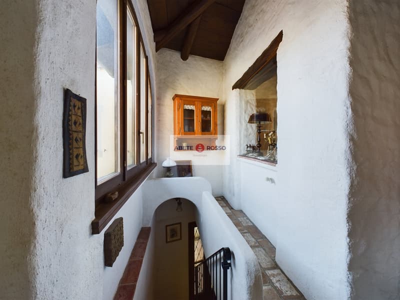 The image shows the interior of a building with a wooden ceiling, white walls, and large windows. There is a wooden cabinet and a lamp visible, as well as some artwork on the walls. The space appears to be a hallway or entryway with a balcony or terrace v