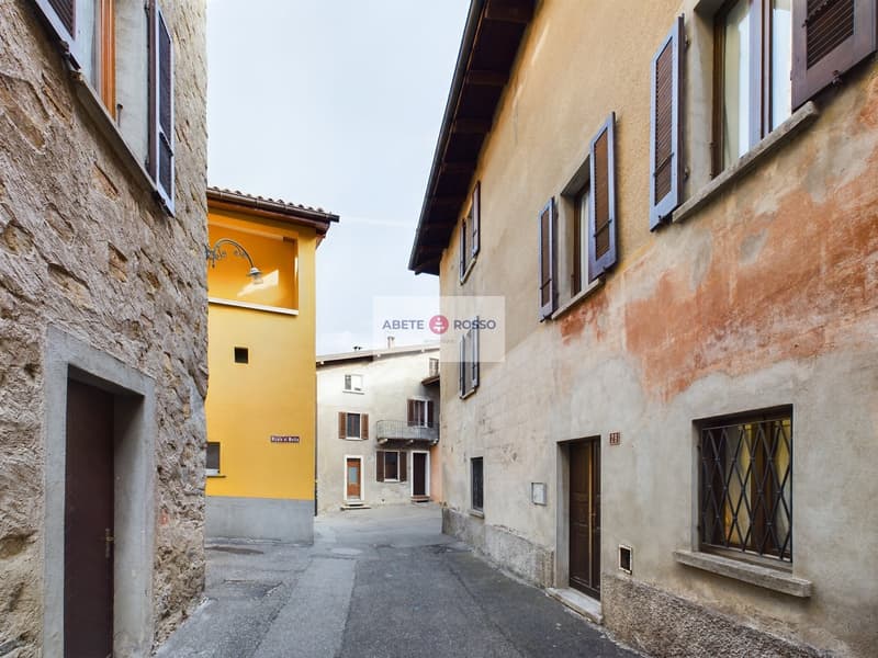 Narrow alley with stone and stucco buildings, some with yellow facades, wooden shutters, and a sign for 'Abete Rosso' on the wall.
