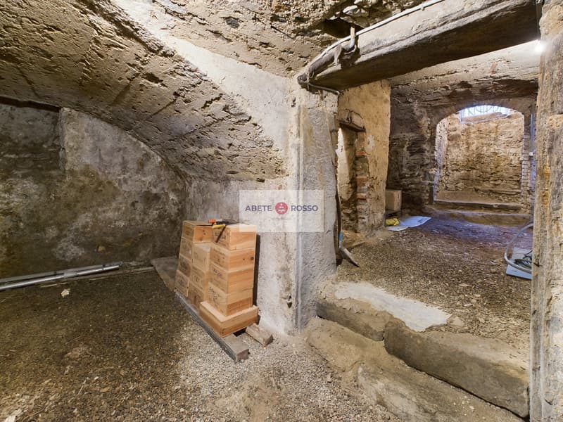 The image shows an underground space with rough stone walls and a vaulted ceiling. There are wooden crates and boxes stacked in the corner, and a sign that says 'Abete Rosso' which appears to be a brand or organization name. The space has a utilitarian, s