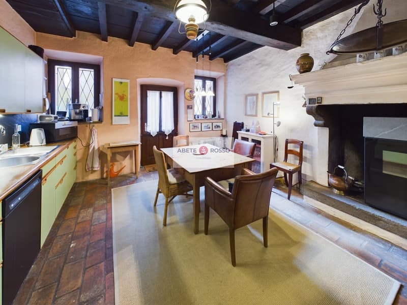 This image shows a rustic and cozy kitchen and dining area. The room has exposed wooden beams on the ceiling, brick floors, and a fireplace. The kitchen features a green vintage-style appliance and a wooden dining table with chairs. The walls are decorate