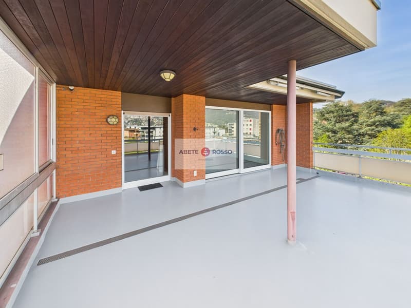 The image shows the exterior of a building with a wooden ceiling and brick walls. The building has large windows and a glass door with the 'ABETE ROSSO' logo displayed. The entrance area has a concrete floor and a railing. The surrounding area appears to