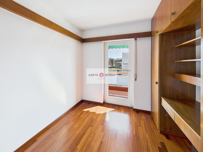 Spacious bedroom with hardwood floors, white walls, and wooden built-in shelving units. The room features a glass door leading to a balcony or terrace, providing natural light and access to an outdoor space.