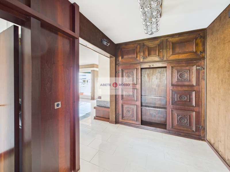 The image shows a spacious entryway with a large, ornate wooden cabinet or wardrobe. The cabinet features intricate carved designs and a warm, rustic finish. The floor is tiled, and there is a chandelier hanging from the ceiling, adding a touch of eleganc