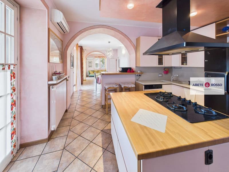 Contemporary style kitchen with wooden countertops, gas stove, pink walls, white ceiling, arched passageway, white cabinets, chandelier, two stools, white radiator, and framed artwork