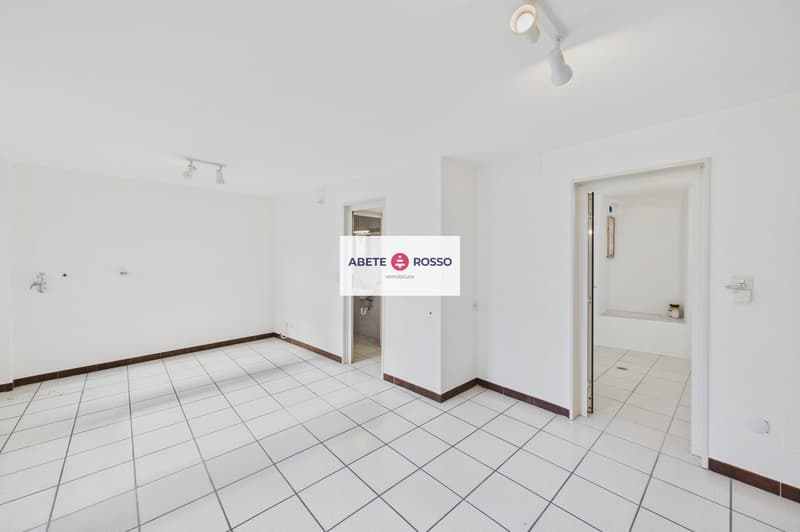 Empty room with white tiles, white walls, ceiling lights, open door, and a small sink area