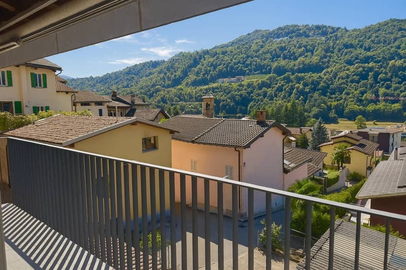 Multi-story apartment building with tiled roofs, surrounded by lush green hills and mountains in the background. The building has balconies with metal railings, providing a scenic view of the natural landscape.