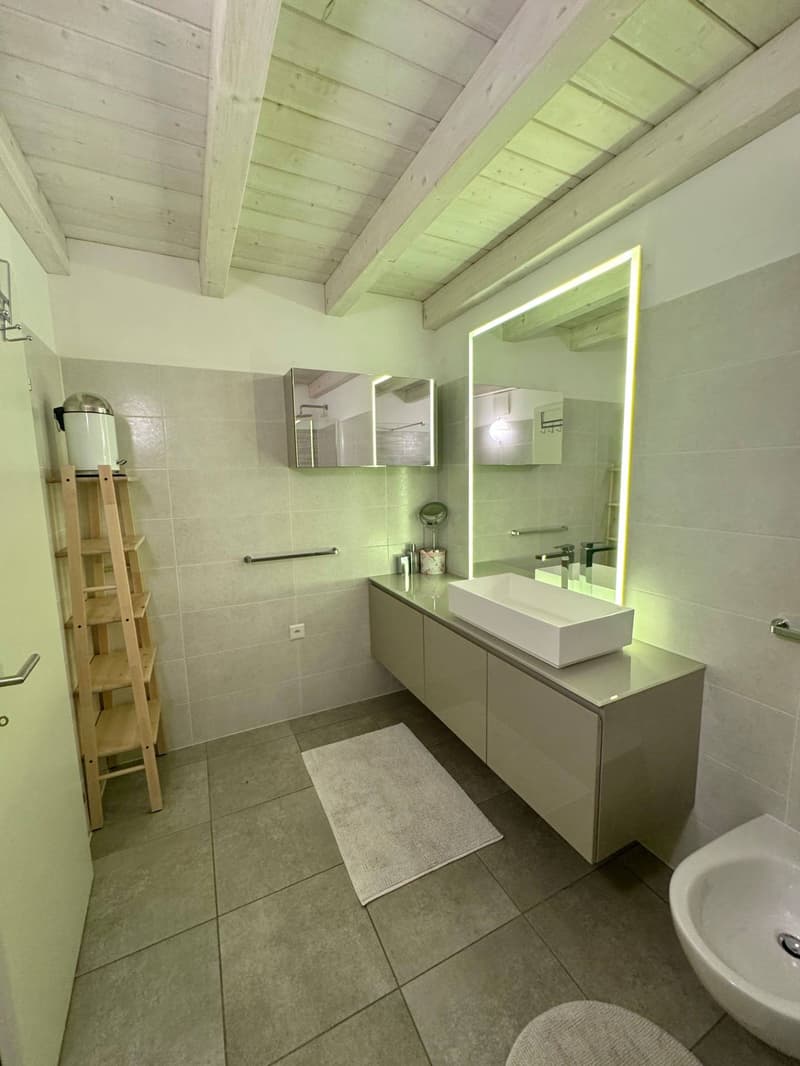 Spacious bathroom with wooden ceiling beams, gray tile flooring, vanity with sink, mirror with lighting, and a ladder shelf unit.
