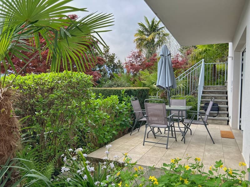 The image shows a well-landscaped outdoor patio area with a variety of lush, tropical plants and greenery. There is a patio set with a table and chairs, as well as a closed umbrella. The patio is surrounded by a railing and steps leading up to the space.