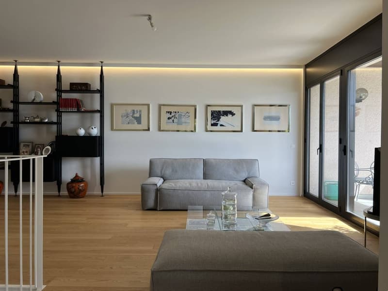 Spacious living room with hardwood floors, gray sofa, glass coffee table, and framed artwork on the walls. The room has large windows that provide natural light and access to a balcony or terrace.