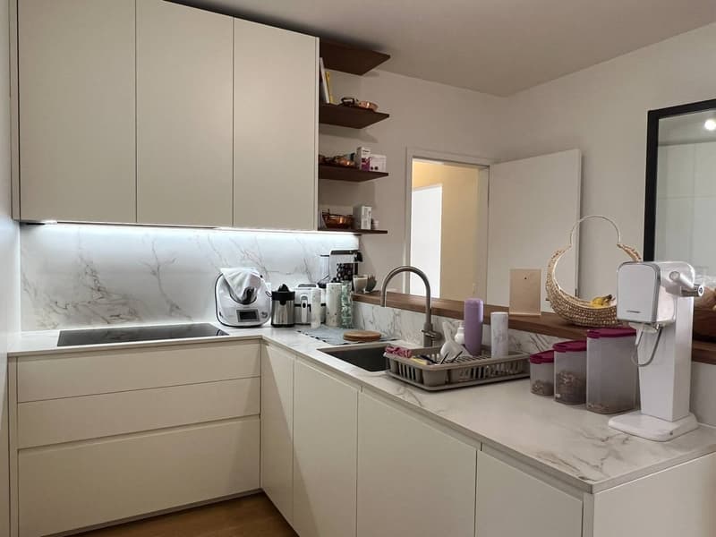 Modern kitchen with light-colored cabinets, marble-like countertops, open shelving, and various kitchen appliances and accessories such as a coffee maker, toaster, and decorative items.