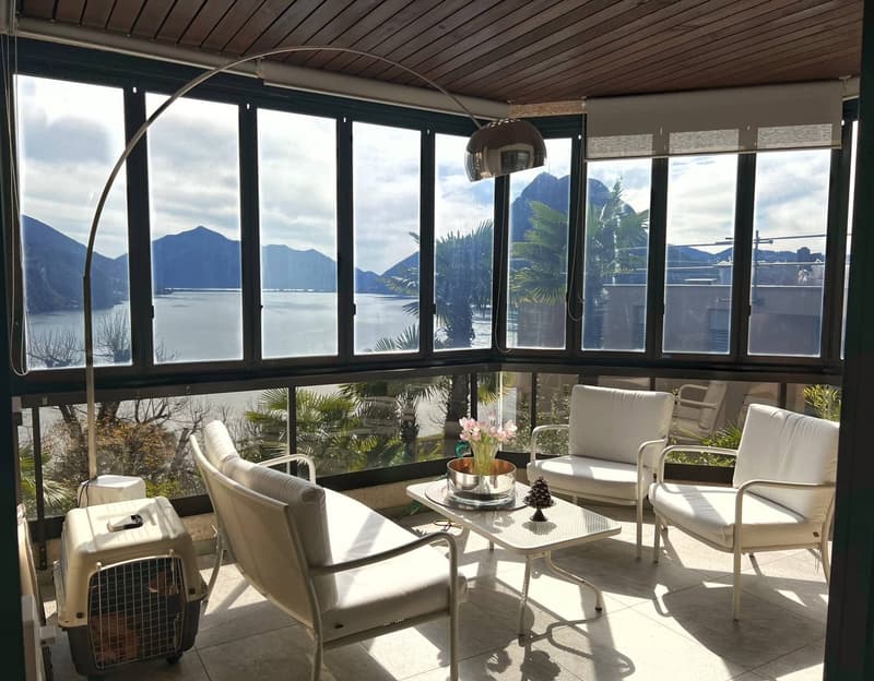 Spacious living room with large windows offering a panoramic view of the mountains and lake. The room features comfortable white sofas and chairs, a coffee table, and a potted plant. The ceiling has a wooden design and there is a heating/cooling unit visi