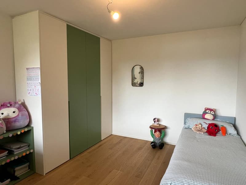 The image shows a bedroom with a white wall, a large wardrobe with green doors, a bed with a striped bedsheet, and various toys and decorations such as a plush animal and a figurine on a shelf.