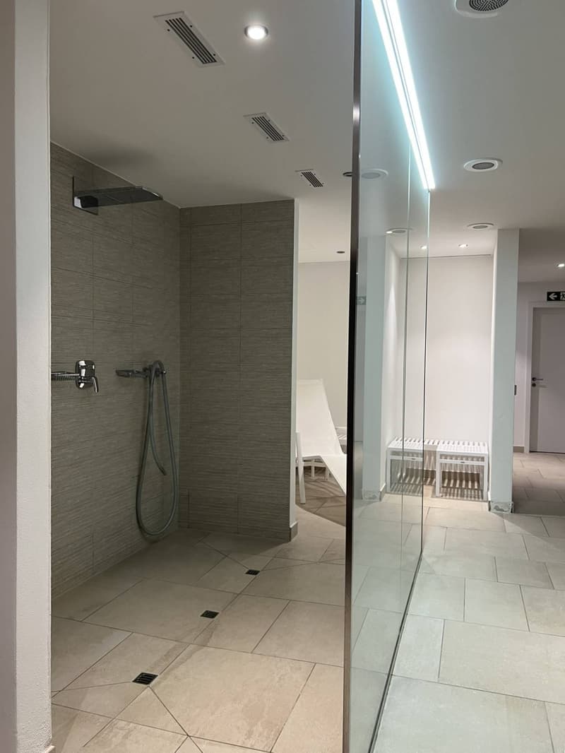 Shower area with grey tiles, glass wall, silver faucet, floor drains, white lounge chairs