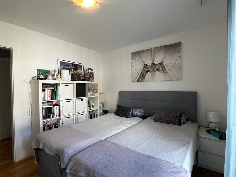 Spacious bedroom with two single beds, white linen, wooden shelves with books and decorative items, night tables with lamps, and picture frames on the wall.