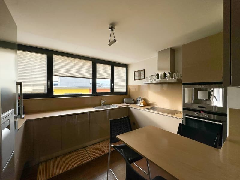 Modern kitchen with dark cabinets, stainless steel appliances including a dishwasher, large windows, and a dining table with chairs