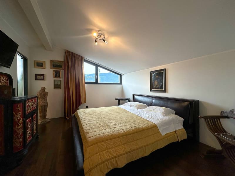 Spacious bedroom with large windows offering a scenic view, a king-size bed with a yellow quilted comforter, a leather headboard, and artwork on the walls. The room also features a TV and other decorative elements.