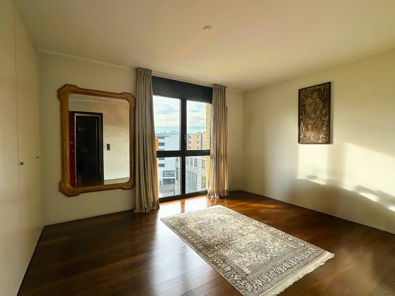 Spacious bedroom with hardwood floors, large windows providing a view, a decorative mirror on the wall, and a patterned area rug on the floor.