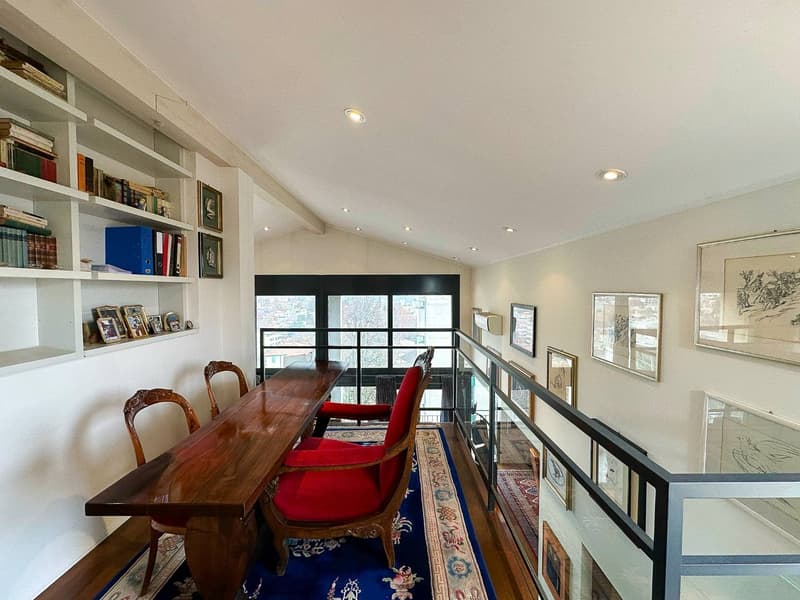 Spacious living room with large windows, wooden dining table, red upholstered chairs, built-in bookshelves, and framed artwork on the walls.