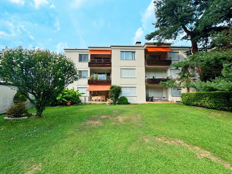Two story apartment building, beige paint, awnings, large front yard with green grass, small garden with trees, street side parking