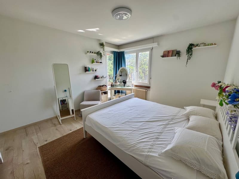 Spacious bedroom with white walls and wooden floor. Includes a bed with white bedspread and pillows, a chair, a mirror, a desk with a lamp, a heater, and shelves with various items. An open window offers a view of trees outside.