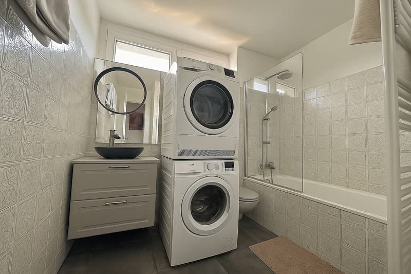 tiled walls, white cabinet, bathtub, shower, toilet, stacked washer and dryer, sink, towel racks